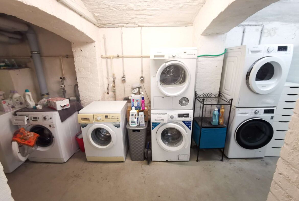 Row of front-loading washing machines and dryers in a basement laundry room with exposed pipes along white brick walls.