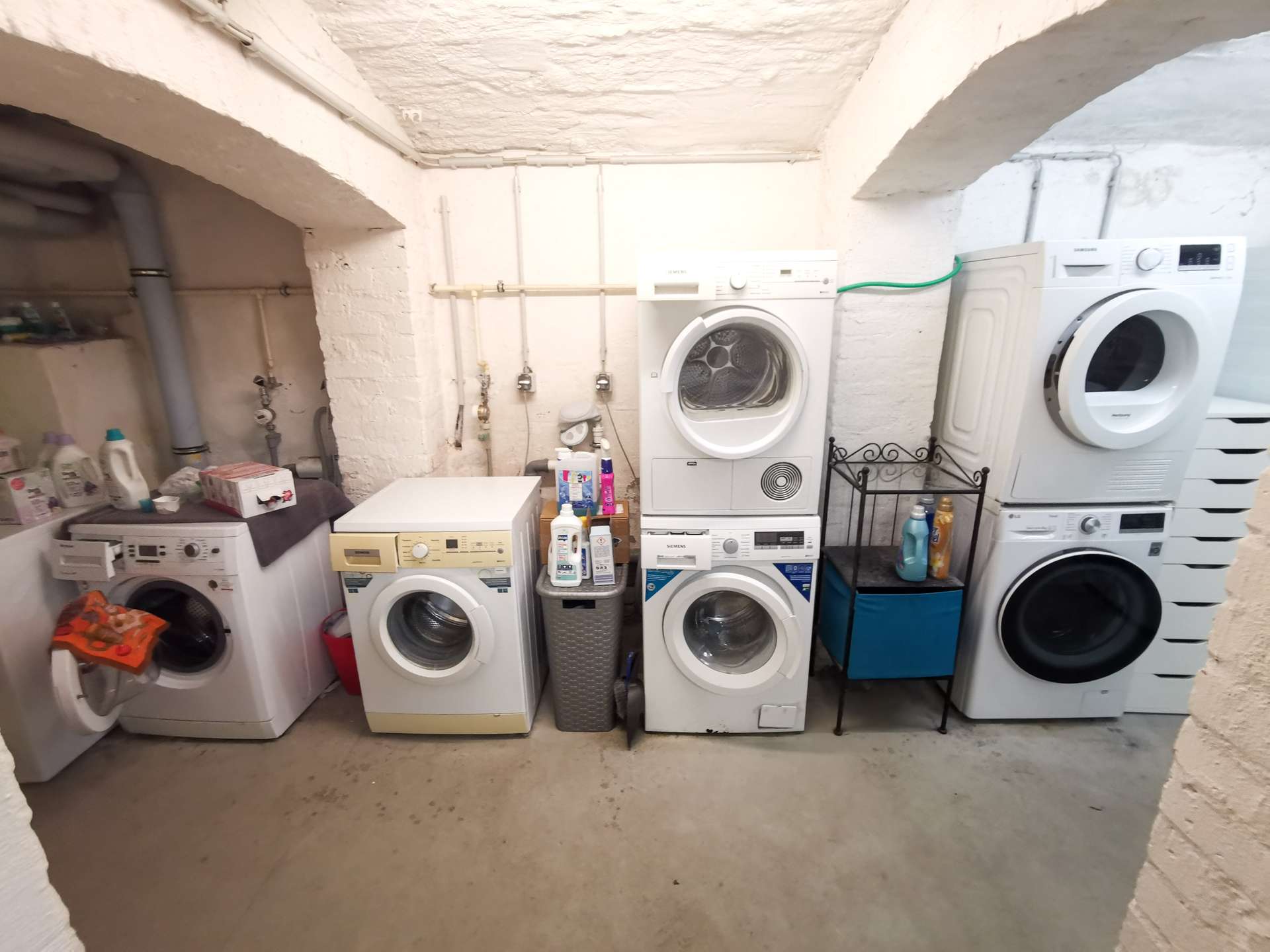 Row of front-loading washing machines and dryers in a basement laundry room with exposed pipes along white brick walls.