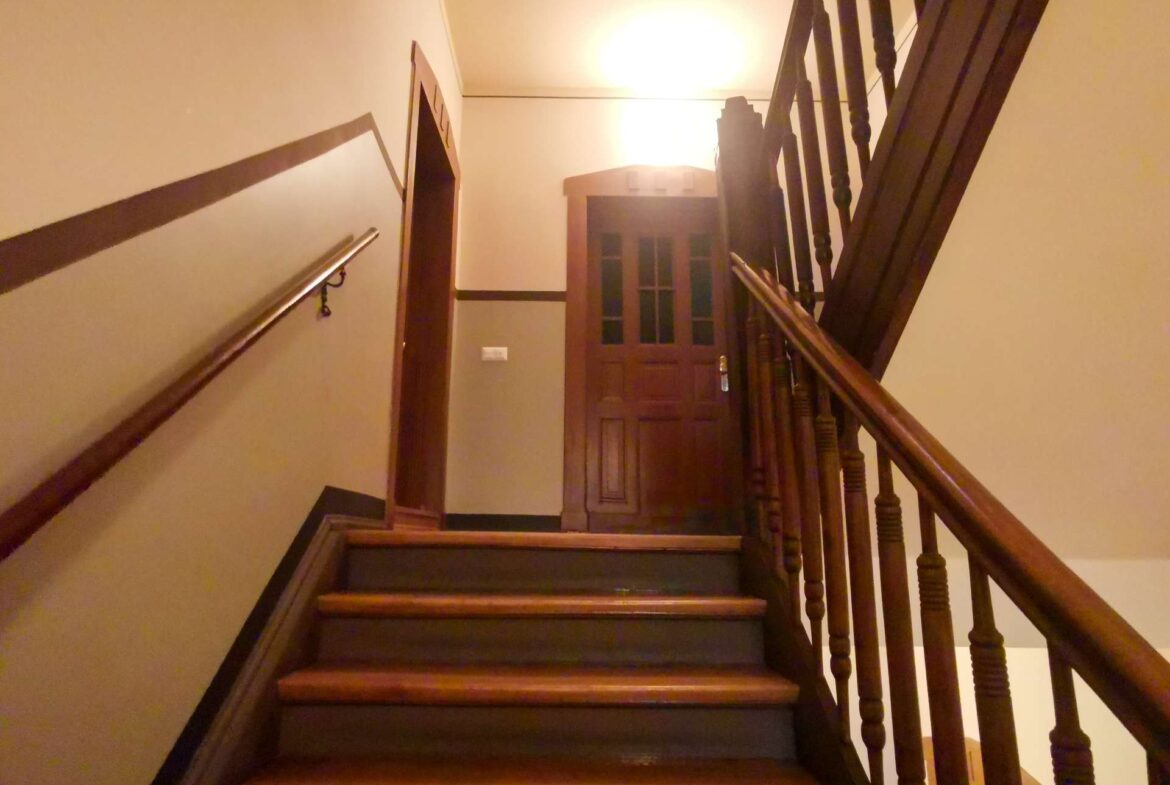 Wooden staircase with ornate balusters leads up to a closed wooden door under warm overhead lighting.