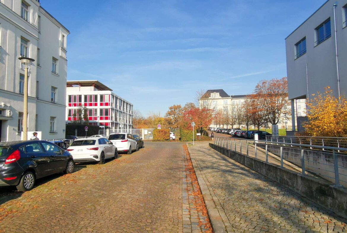Cobblestone street lined with parked cars and white buildings, autumn trees with orange leaves under a clear blue sky.