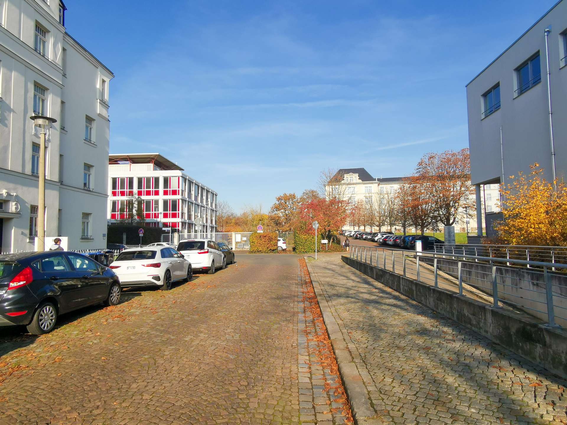 Cobblestone street lined with parked cars and white buildings, autumn trees with orange leaves under a clear blue sky.