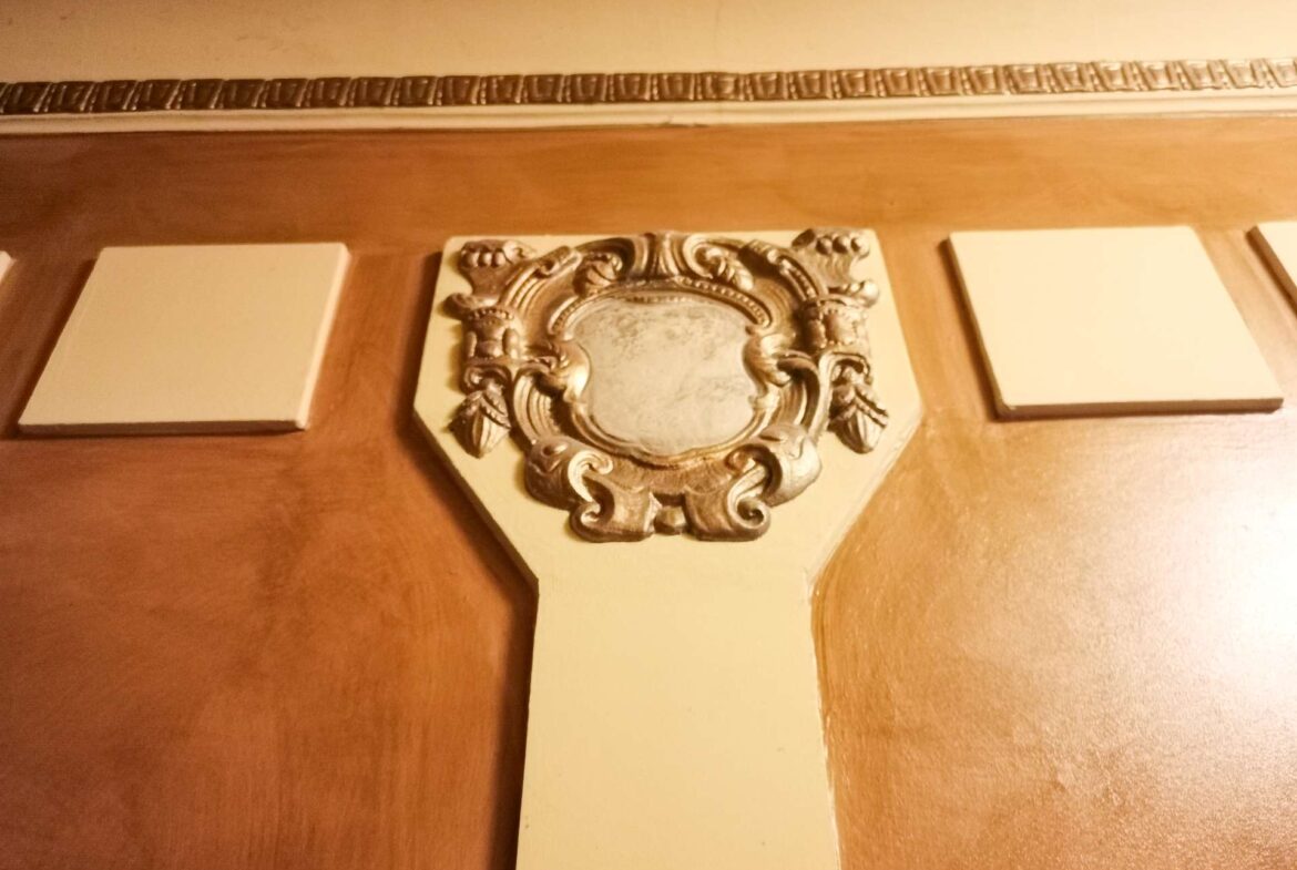 Decorative wooden cross with an ornate silver central emblem and three blank white plaques along the arms on a brown wall.