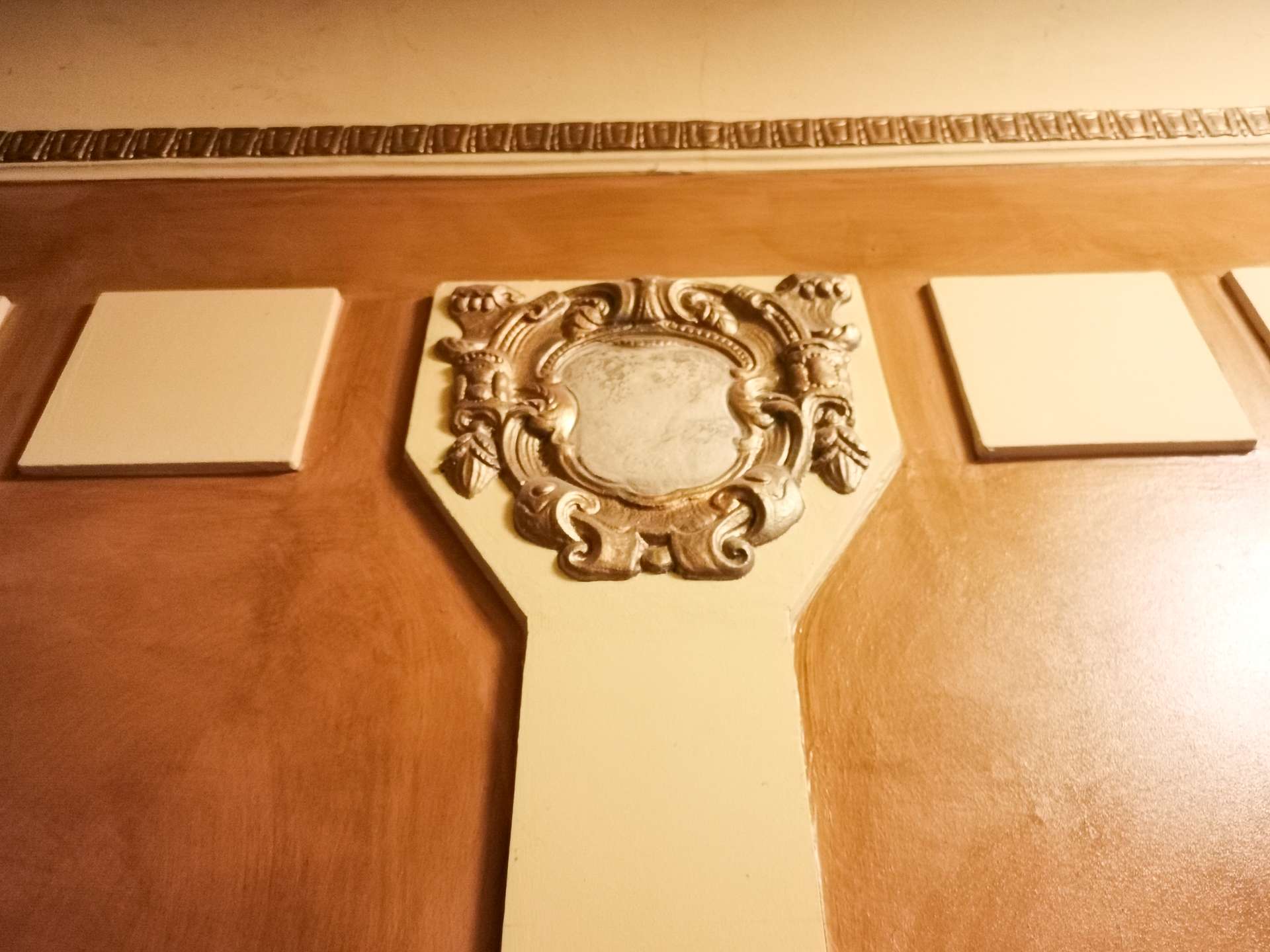 Decorative wooden cross with an ornate silver central emblem and three blank white plaques along the arms on a brown wall.