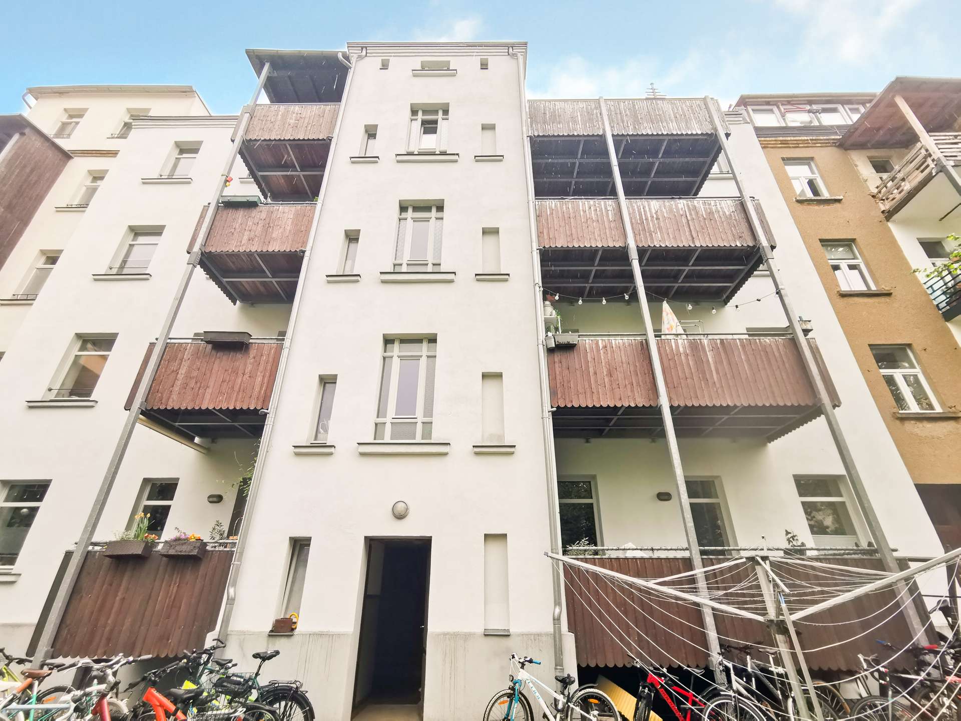 White multi-story residential building with brown wooden balconies, a central entry, and bicycles parked along the ground in front of the courtyard.