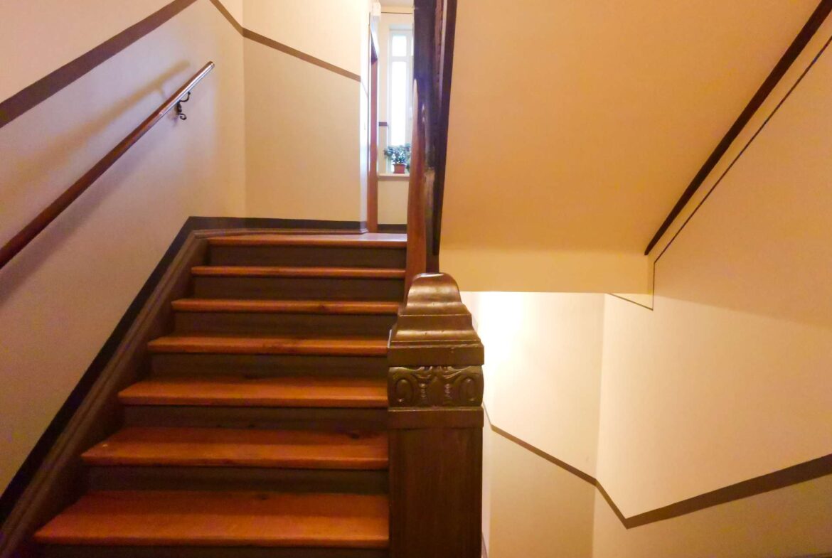 Wooden staircase in a narrow hallway, beige walls with dark brown trim and a carved newel post at the bottom left. A window with a small plant is visible at the top.