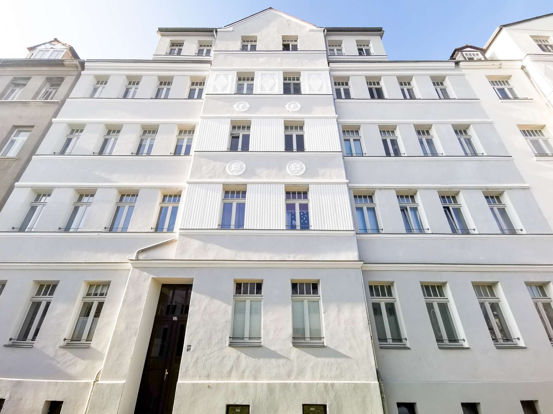 White multi-story apartment building viewed from ground level, rows of windows and pale facade under a clear blue sky.