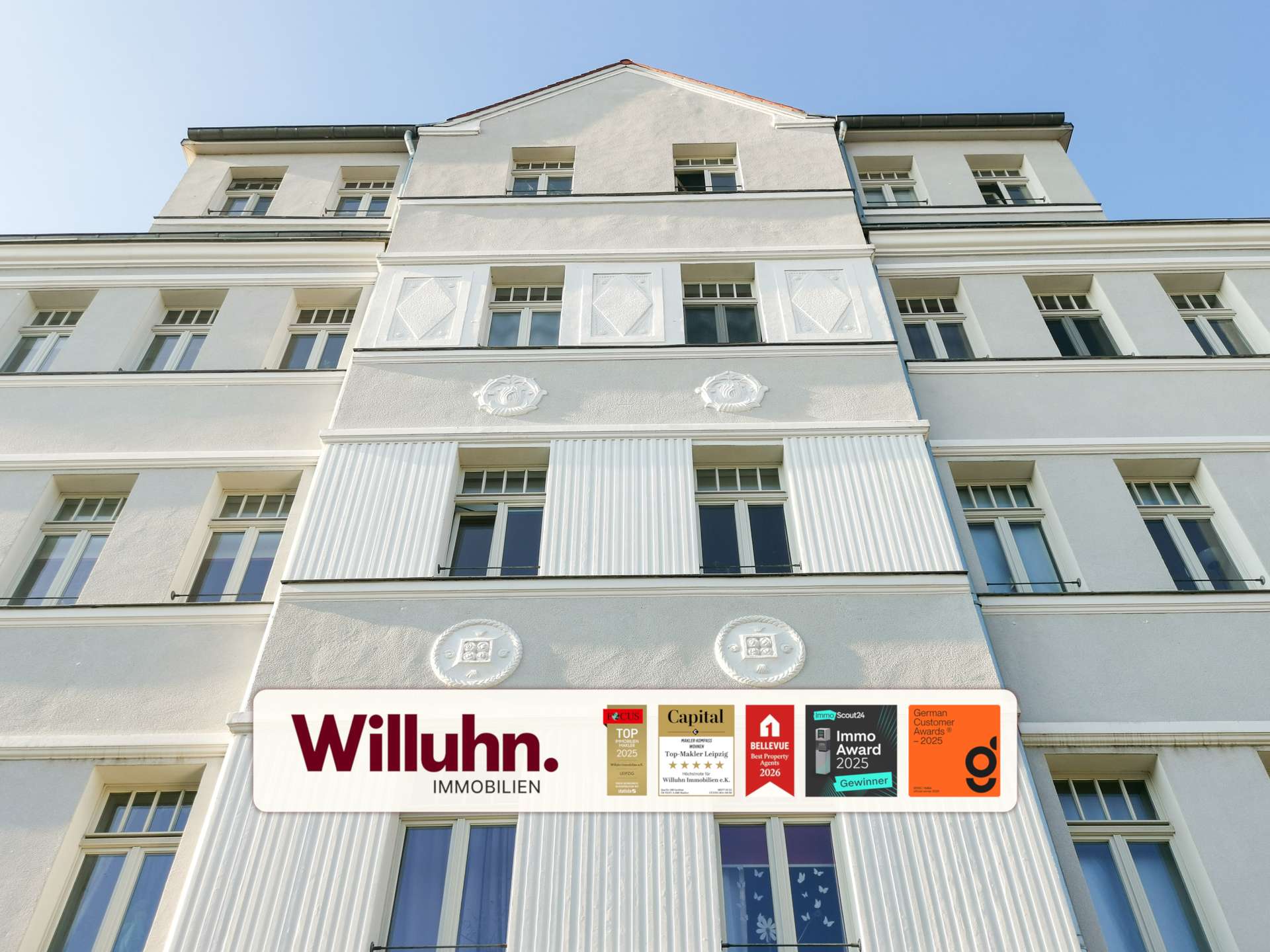 White multi-story building facade with a large Willuhn Immobilien sign and award stickers at street level under a blue sky.