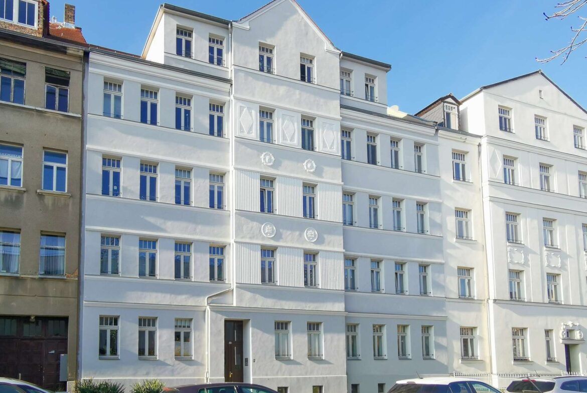 White multi-story residential building with many windows and decorative details, parked cars along the street in front under a clear blue sky