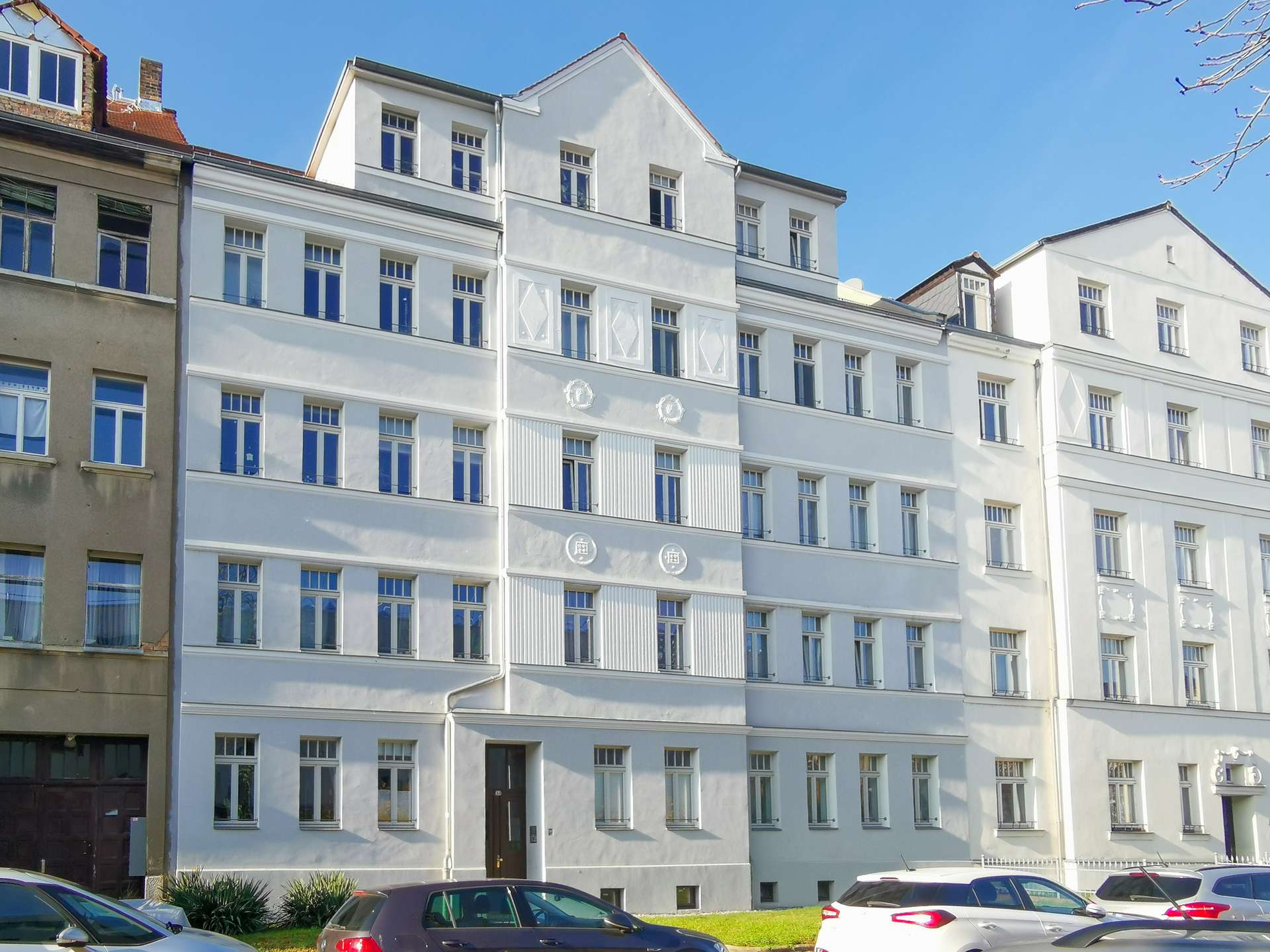 White multi-story residential building with many windows and decorative details, parked cars along the street in front under a clear blue sky