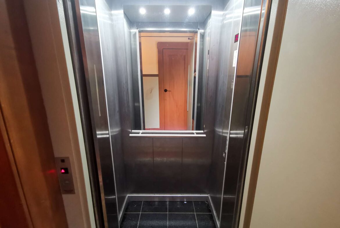 Open elevator cabin with stainless steel walls, dark tiled floor, and a wooden door visible at the far end.