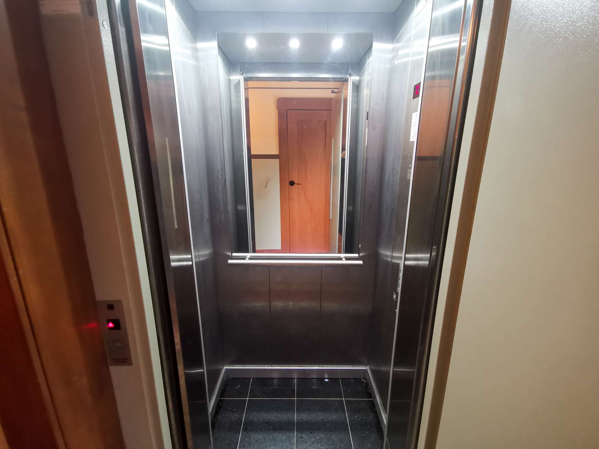 Open elevator cabin with stainless steel walls, dark tiled floor, and a wooden door visible at the far end.