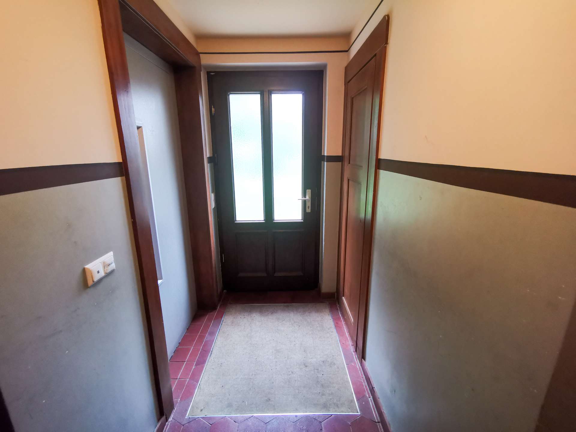 Narrow hallway with brown door frames leading to a dark front door with frosted glass panels; green walls with a dark horizontal stripe and a red tile border by the floor.