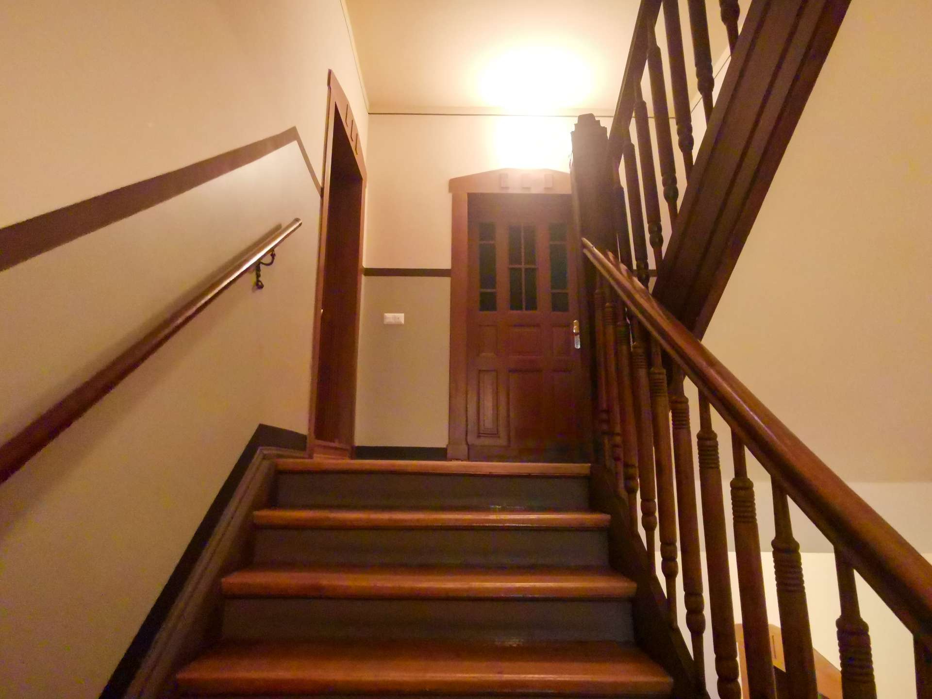 Wooden staircase in a home hallway, with dark railing and balusters, leading up to a wooden door under warm ceiling light.