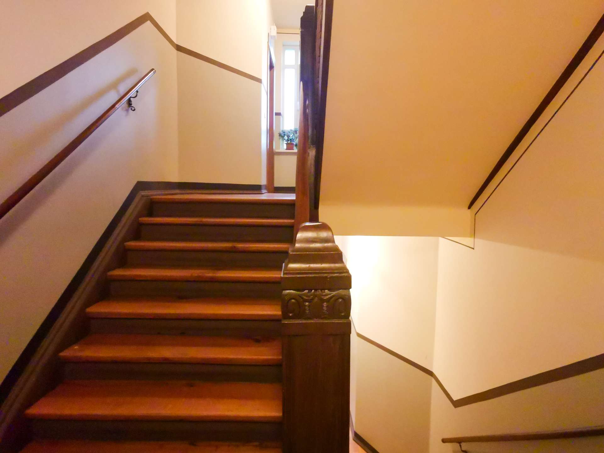 Wooden staircase with carved newel post, beige walls, and dark brown handrails indoors.