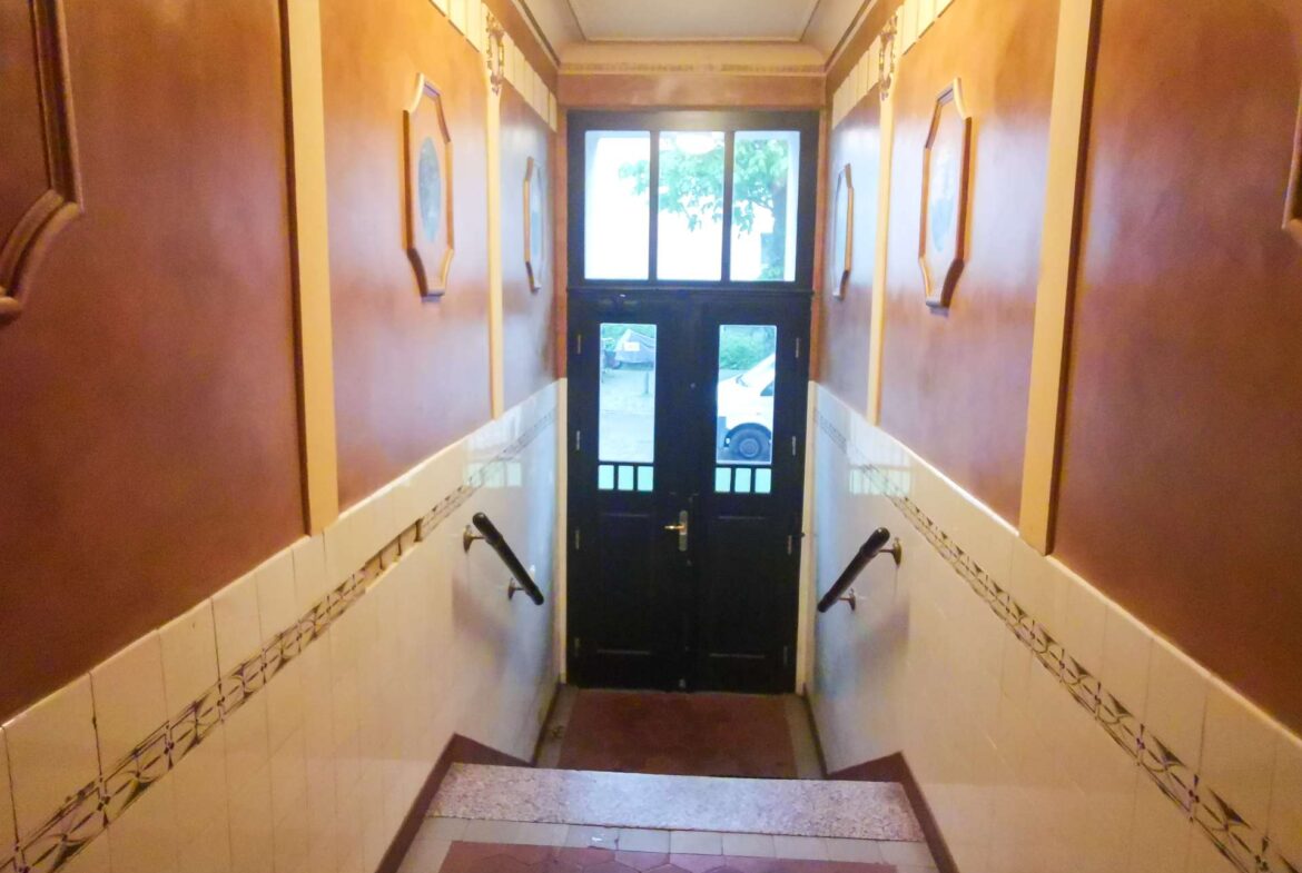 Narrow hallway with brown walls, white tiled lower walls and decorative border, handrails on both sides, ending at a glass‑pane door to the outside.