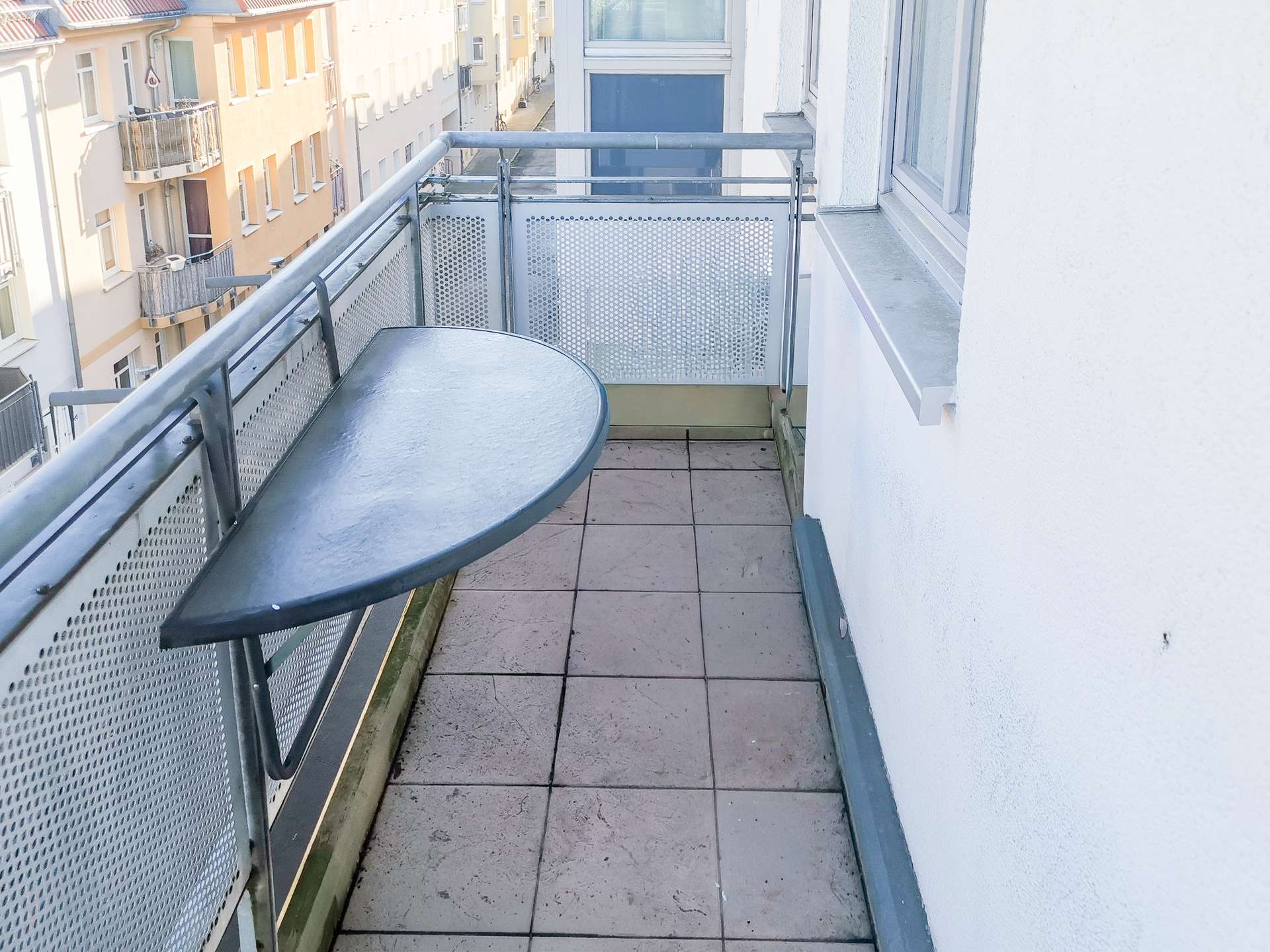 Narrow apartment balcony with a round glass table and metal railing, overlooking a city street with pastel buildings on either side.