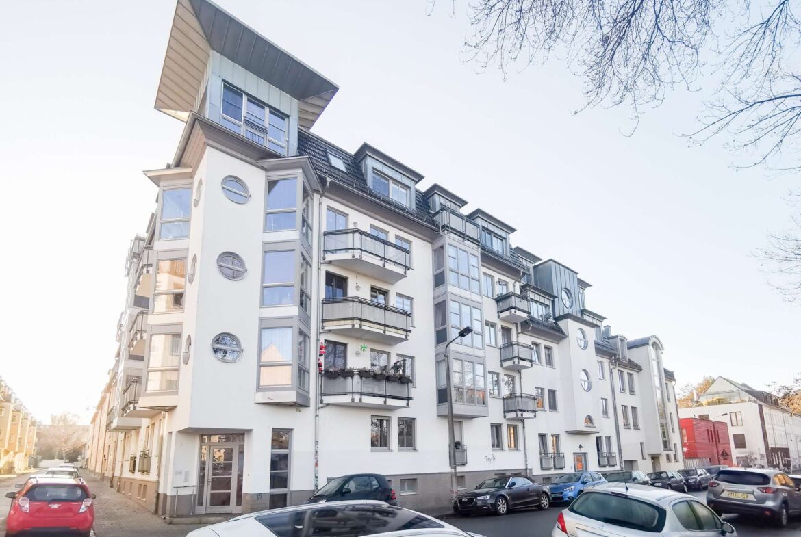 Modern white apartment building with balconies lining a sunlit street and parked cars along the curb.