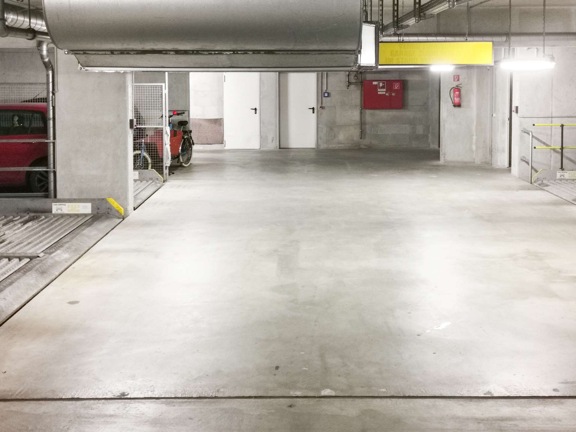 Underground parking garage with a red van on the left, a bike and scooter inside a wire cage, and white doors along the back wall.