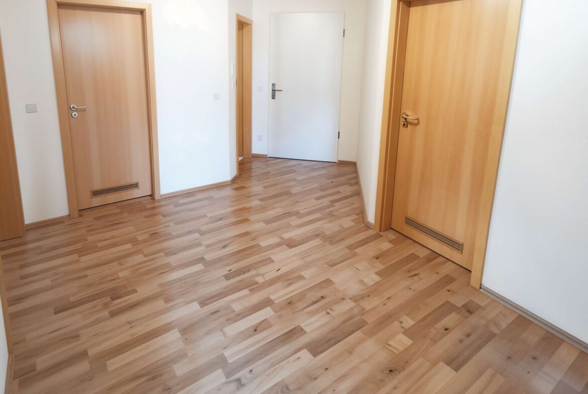 Bright hallway with light hardwood floors and three wooden doors along white walls, seen from a corner perspective.