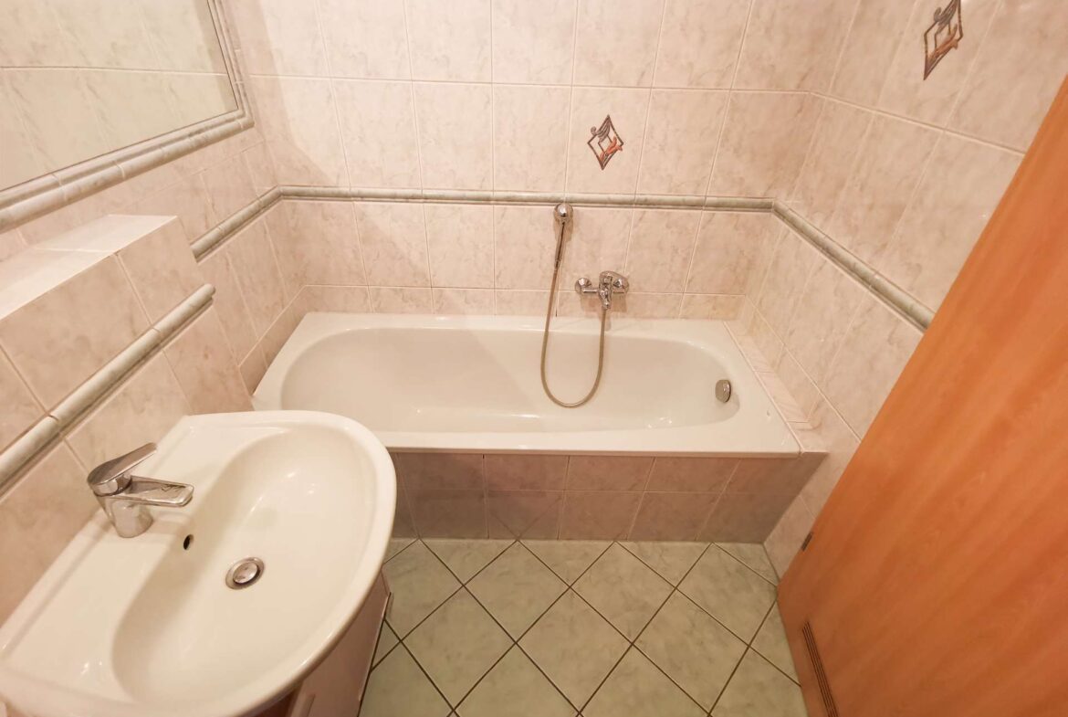 Small beige tiled bathroom with a white bathtub, handheld shower, and a sink with a chrome faucet; mirror on the wall and a wooden door to the right.