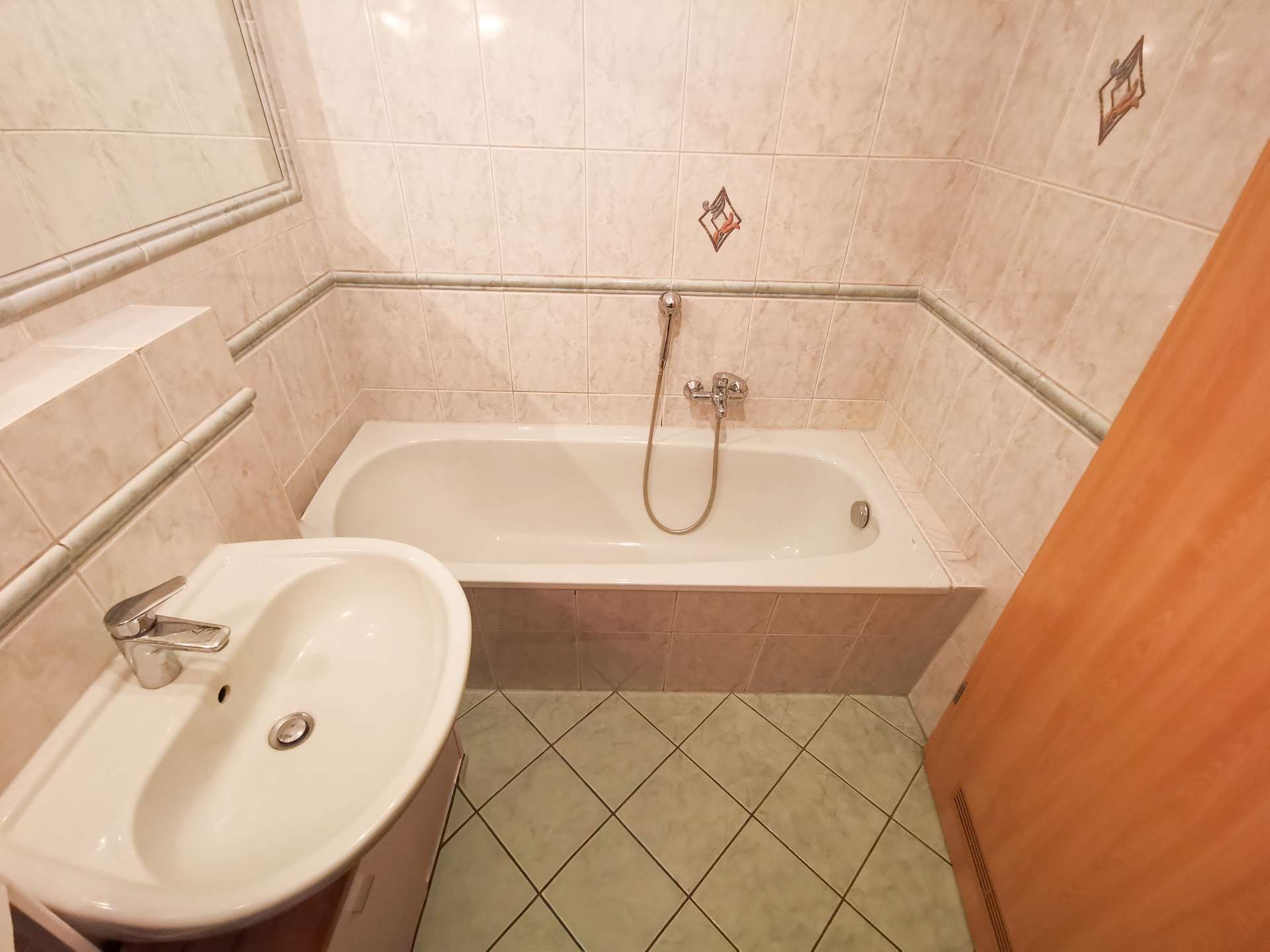 Small beige tiled bathroom with a white bathtub, handheld shower, and a sink with a chrome faucet; mirror on the wall and a wooden door to the right.