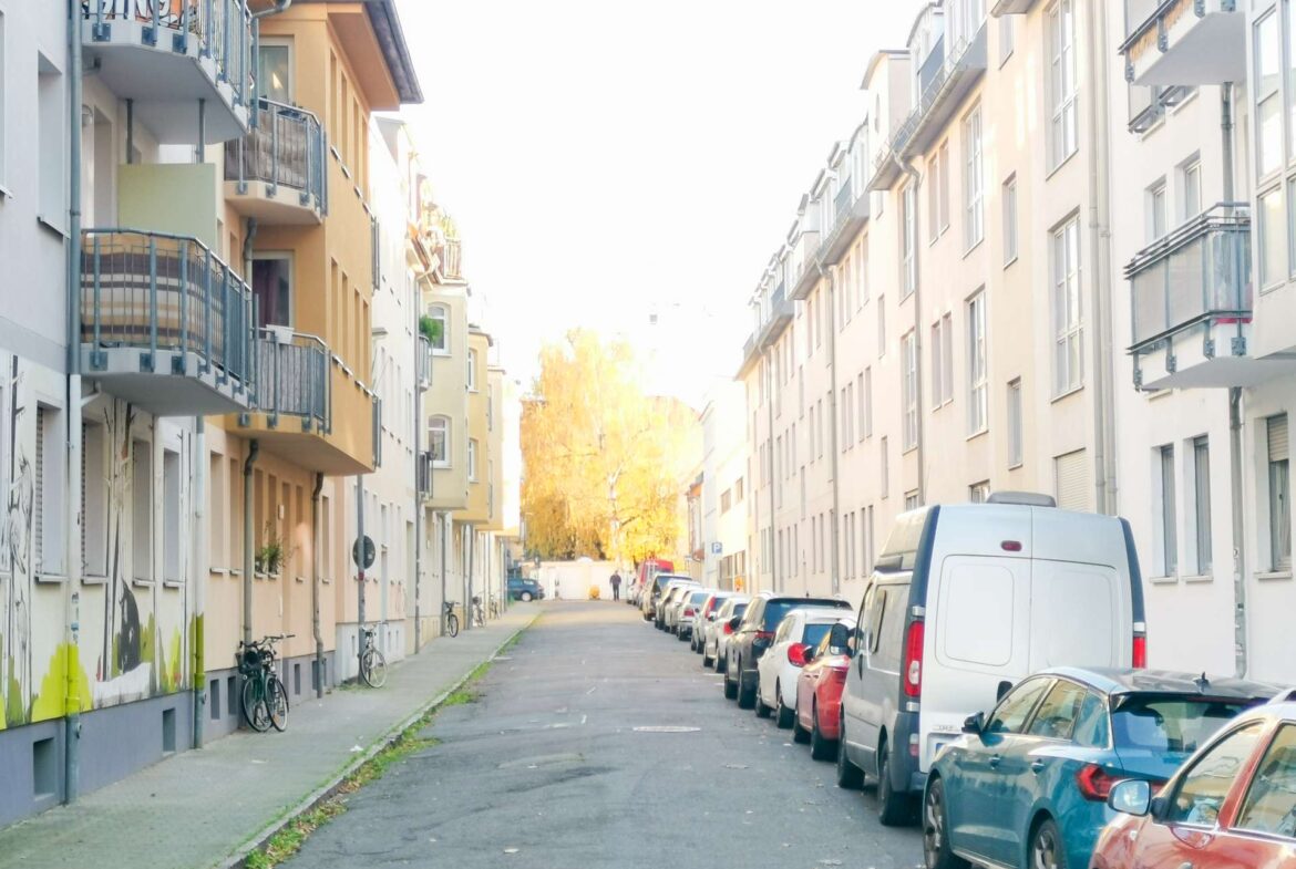 Narrow city street between pastel apartment buildings with balconies, parked cars on both sides, and bicycles along the left sidewalk.