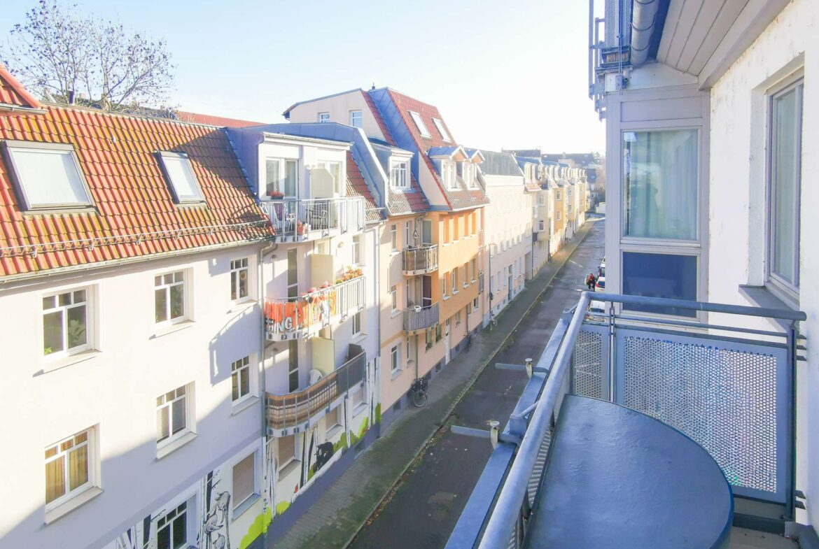 View from a apartment balcony overlooking a sunlit street lined with pastel-colored buildings and red-tiled roofs and small balconies.