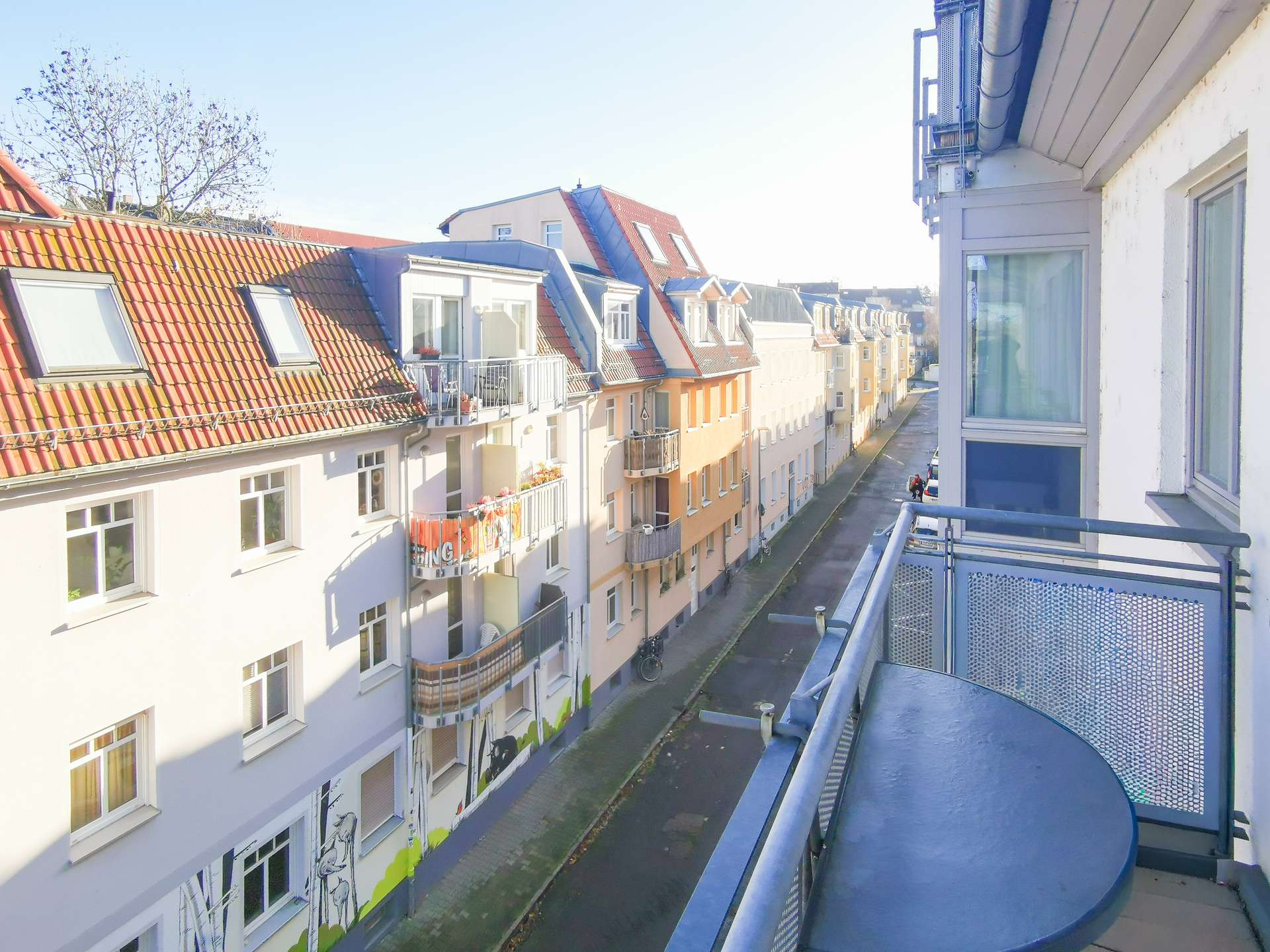 View from a apartment balcony overlooking a sunlit street lined with pastel-colored buildings and red-tiled roofs and small balconies.