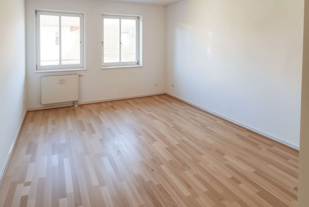 Empty white room with two windows, radiator below left window, and light wood laminate flooring