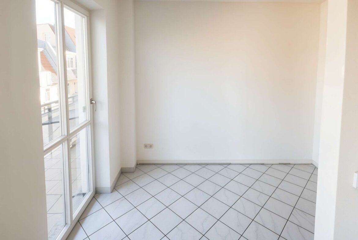 Bright empty room with large glass door/windows on the left leading to a balcony, white walls, and tiled floor in daylight.