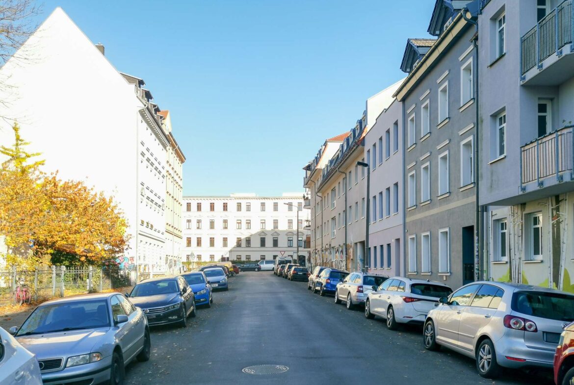 Residential street in a city with cars parked along both sides and pastel apartment buildings under a clear blue sky.