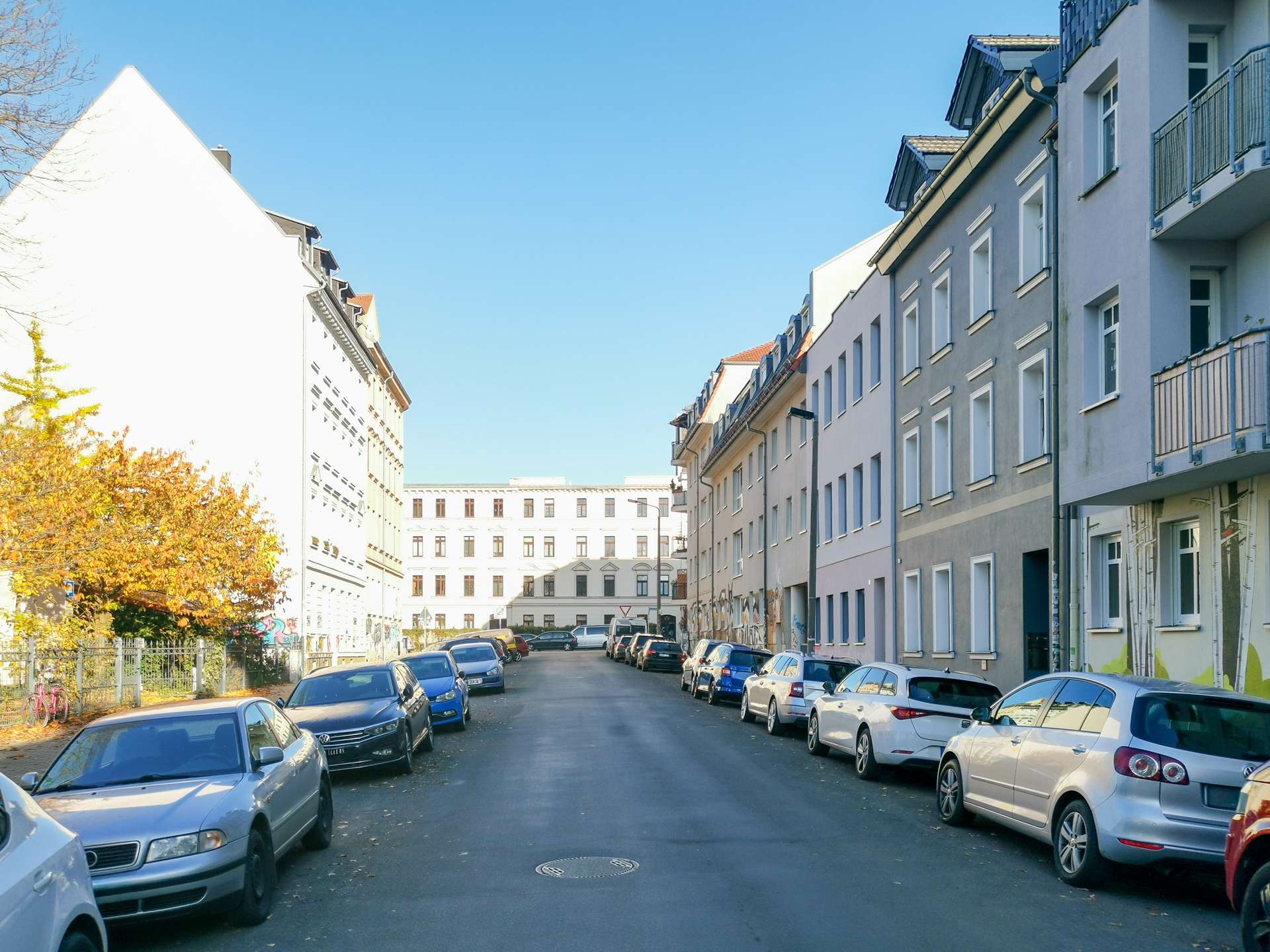 Residential street in a city with cars parked along both sides and pastel apartment buildings under a clear blue sky.