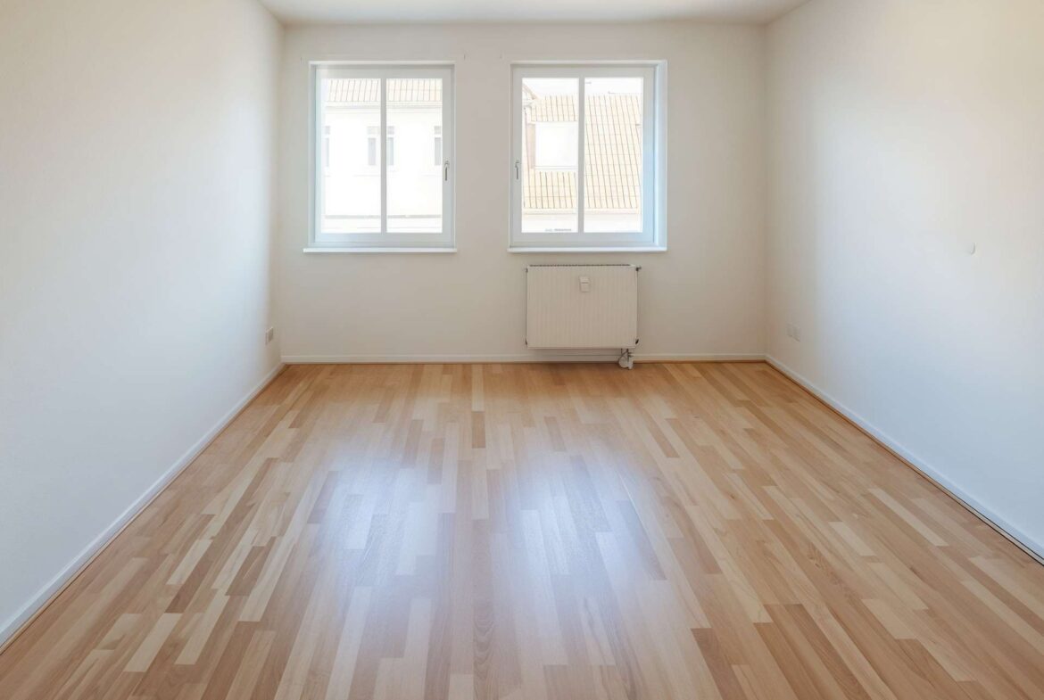 Empty bright room with light wood flooring, two windows on the far wall letting in daylight and a radiator beneath them