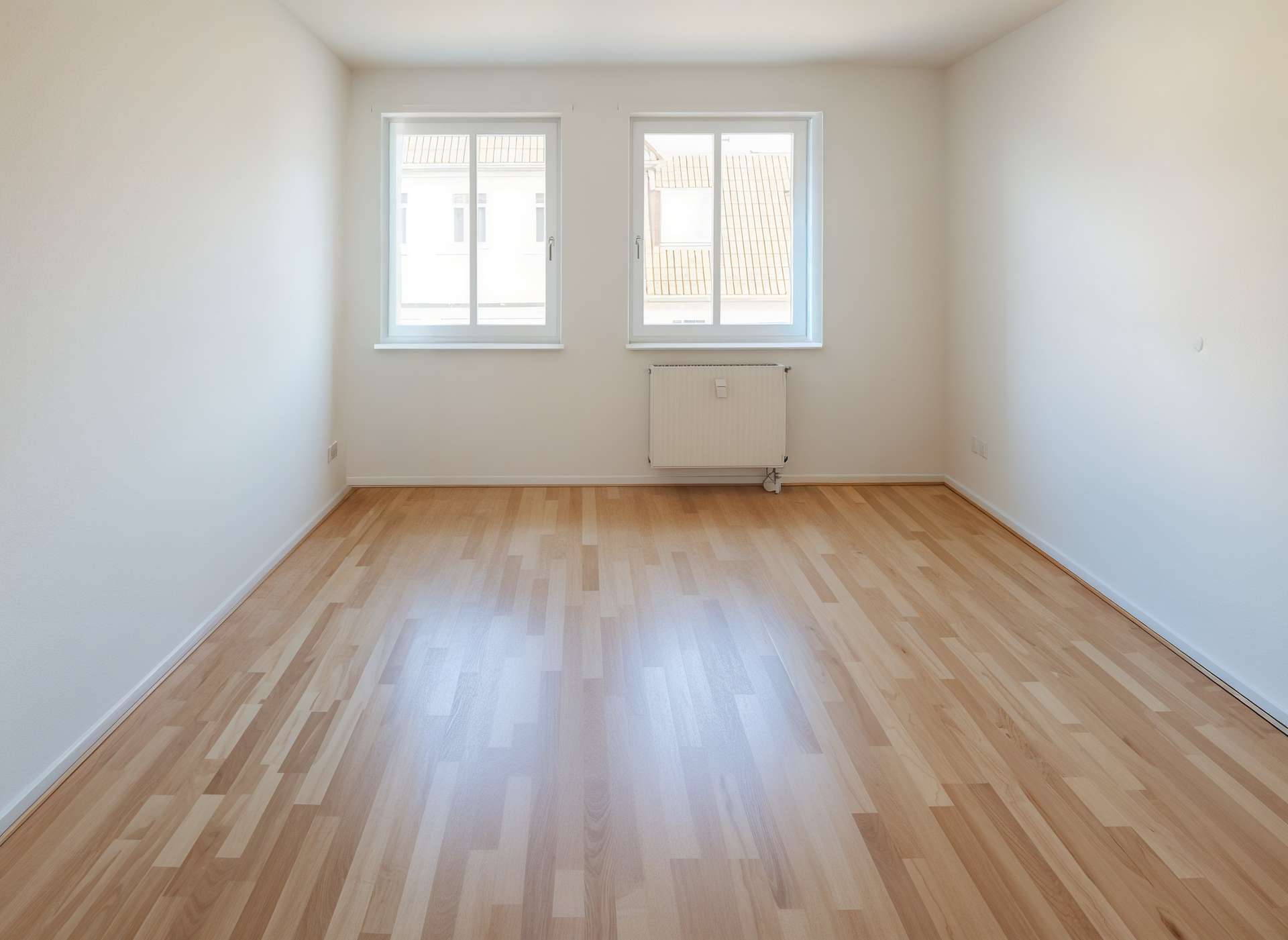Empty bright room with light wood flooring, two windows on the far wall letting in daylight and a radiator beneath them
