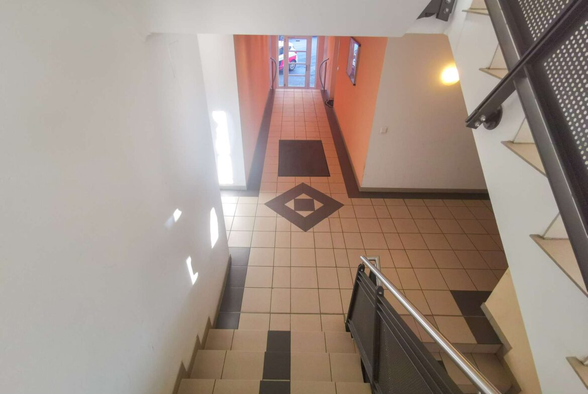 Interior of a stairwell descending to a tiled lobby with orange walls and glass doors at the far end, metal handrails on both sides, and geometric floor pattern.
