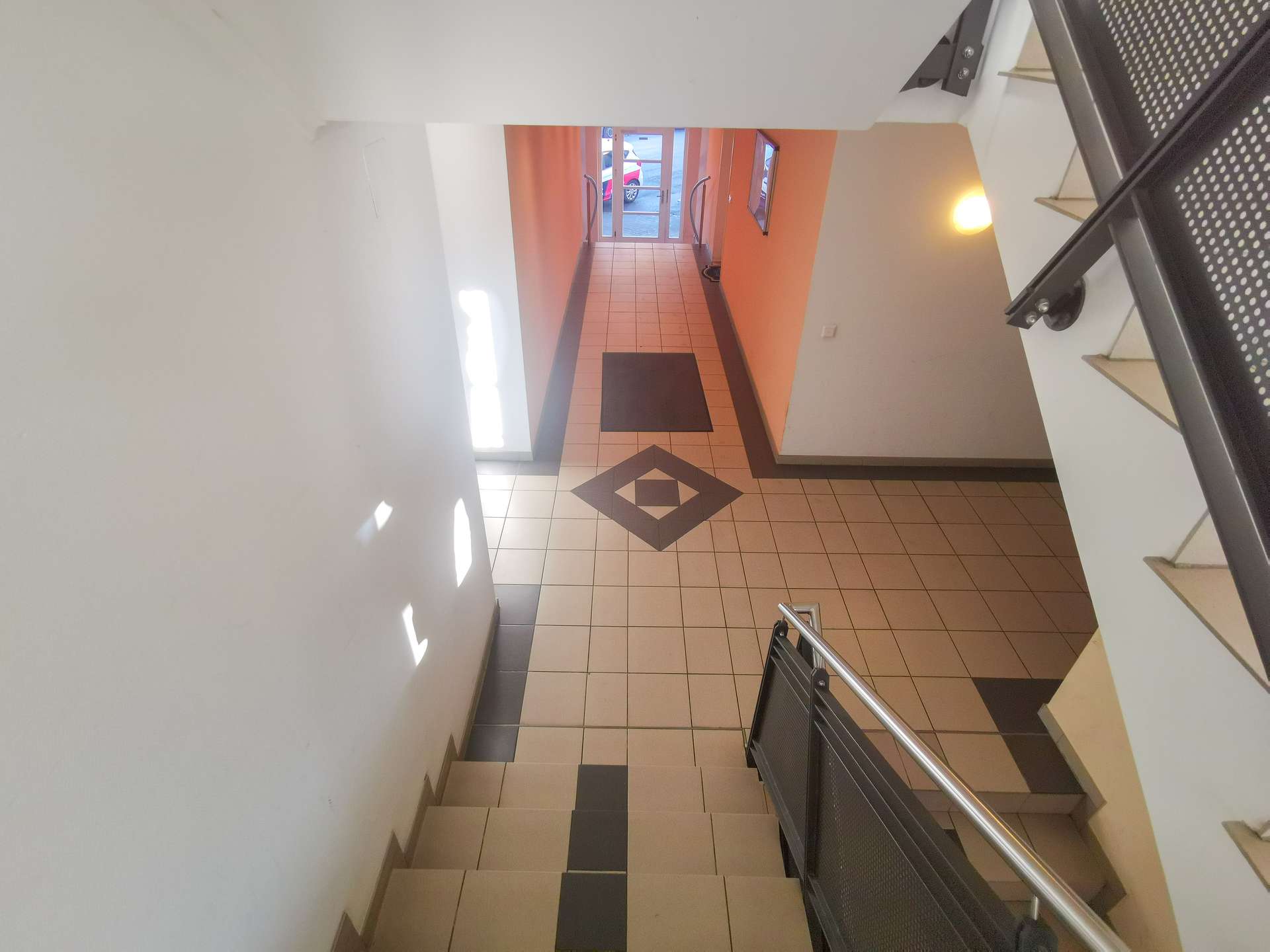 Interior of a stairwell descending to a tiled lobby with orange walls and glass doors at the far end, metal handrails on both sides, and geometric floor pattern.