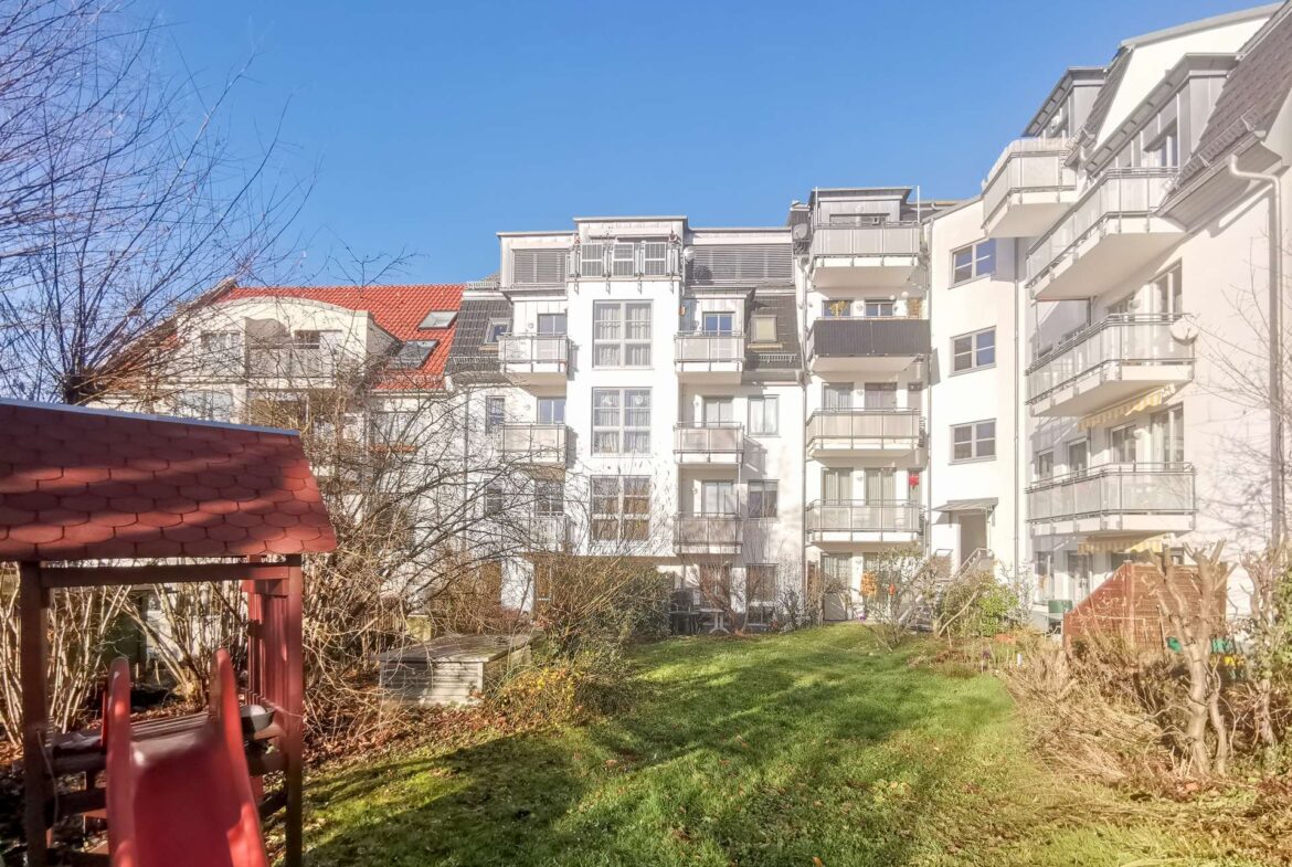 Red playground slide in the foreground with a grassy yard and white apartment buildings with balconies in the background, under a clear blue sky.