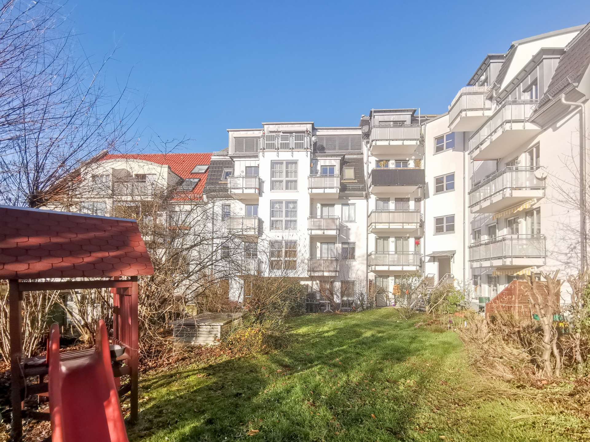 Red playground slide in the foreground with a grassy yard and white apartment buildings with balconies in the background, under a clear blue sky.