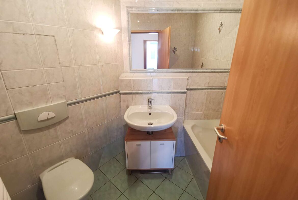 Small tiled bathroom with a wall-mounted toilet, pedestal sink on a wooden cabinet, and a bathtub along the right; door open with a large mirror above the sink reflecting the room.