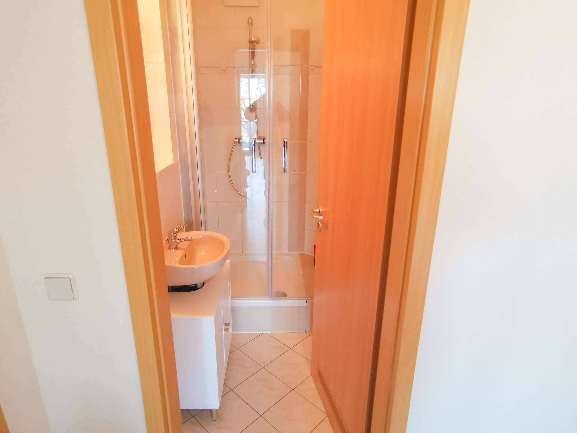 Narrow bathroom seen through a doorway with a glass shower enclosure at the far end and a small sink on the left