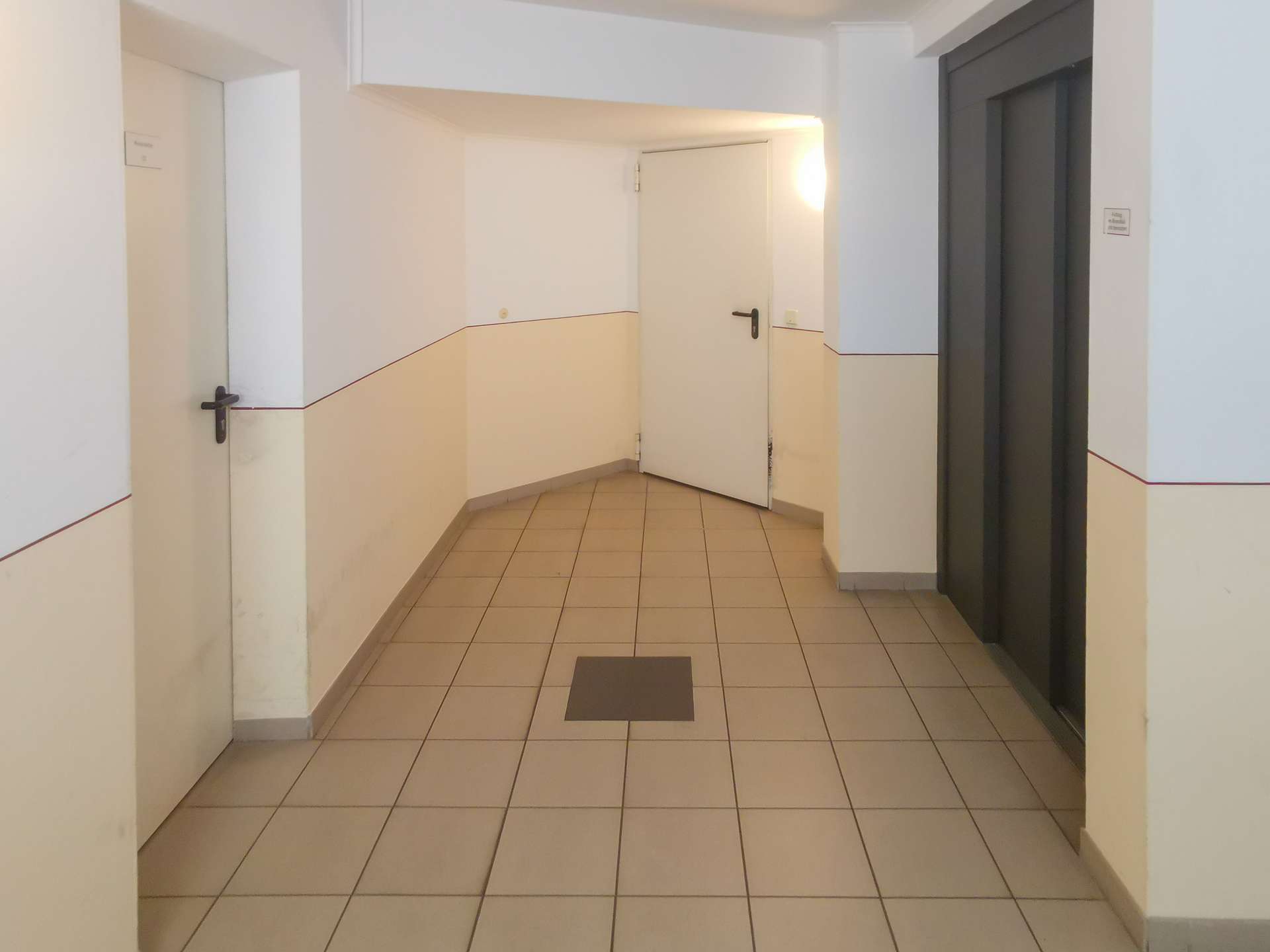 Interior corridor with beige lower walls, white upper walls, and a closed white door at the end; elevator doors on the right; tiled floor