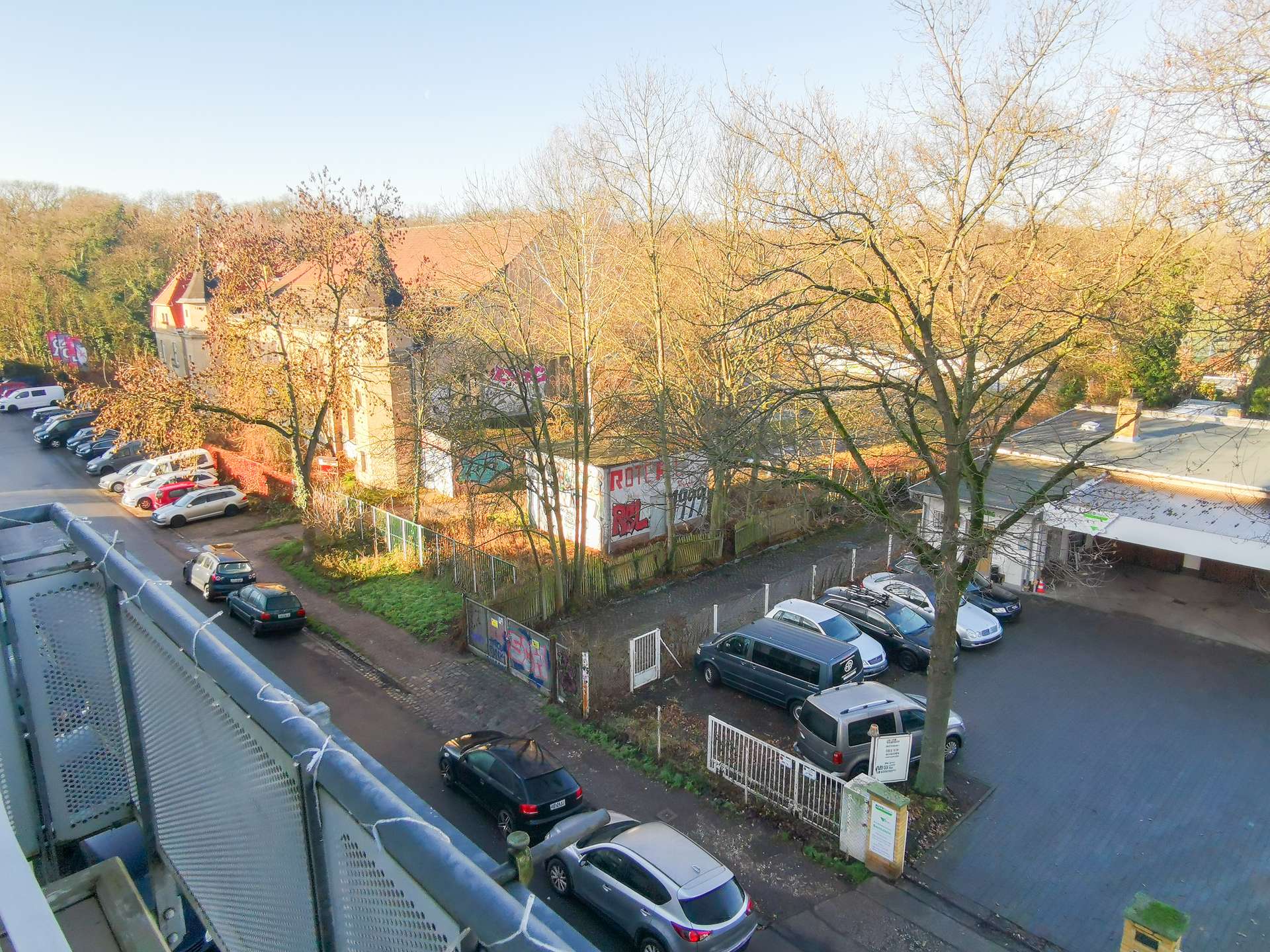Elevated view of a street with many parked cars, leafless trees, and a brick building with graffiti along the sidewalk on a sunny day.