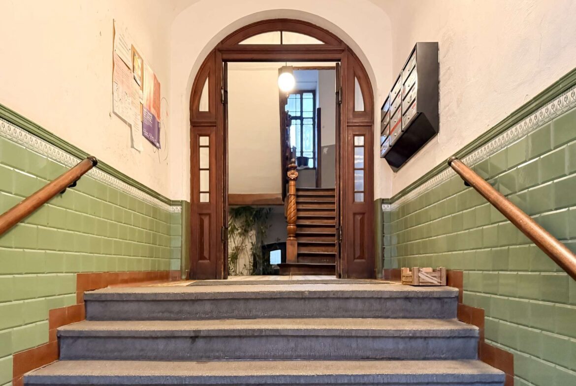 Interior stairwell with green tile wainscoting, wooden arched double doors, and a staircase ascending to a bright upper level; handrails run along both sides.