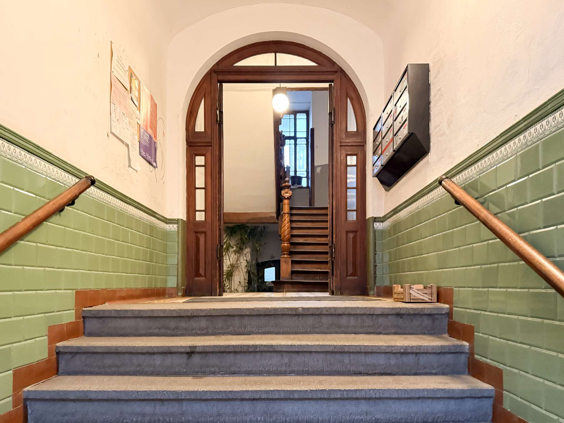 Interior stairwell with green tile wainscoting, wooden arched double doors, and a staircase ascending to a bright upper level; handrails run along both sides.