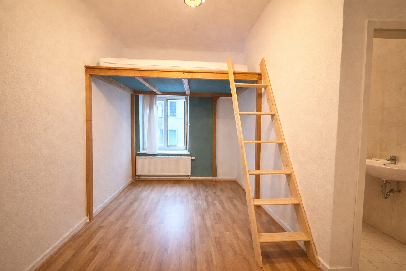 Small studio room with a wooden loft bed, ladder, and a window with radiator beneath; doorway to a tiled bathroom on the right.