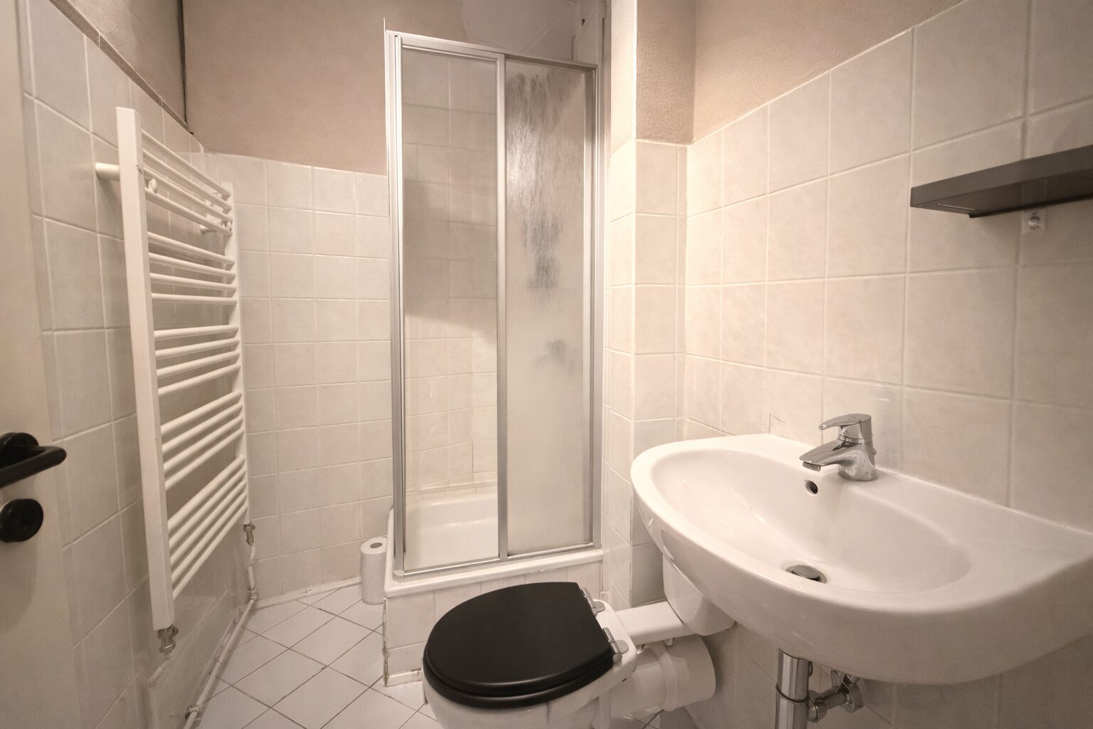 Compact bathroom with shower enclosure, wall tiles, sink with chrome faucet, and a black-seat toilet; white heated towel rail on the left.