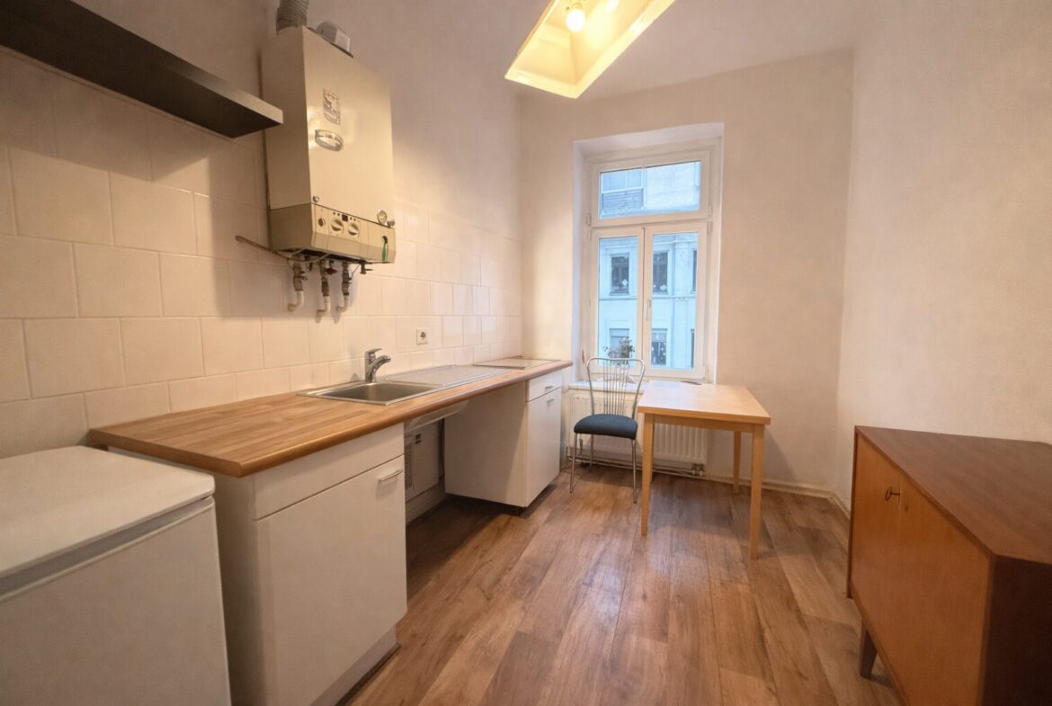 Small kitchen with a wooden countertop, white cabinets, and a dual sink under a window; a wall-mounted boiler above the counter, a metal chair and wooden table near the window, and a wooden sideboard on the right.