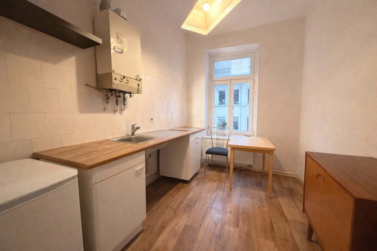 Small kitchen with a wooden countertop, white cabinets, and a dual sink under a window; a wall-mounted boiler above the counter, a metal chair and wooden table near the window, and a wooden sideboard on the right.