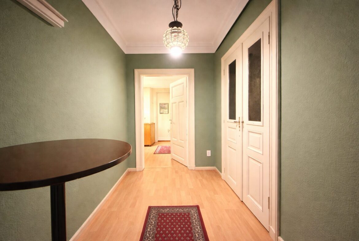 Green-walled hallway with a round wooden table on the left and white double doors on the right, lit by a hanging ceiling lamp; open doorway ahead reveals another room and rug.