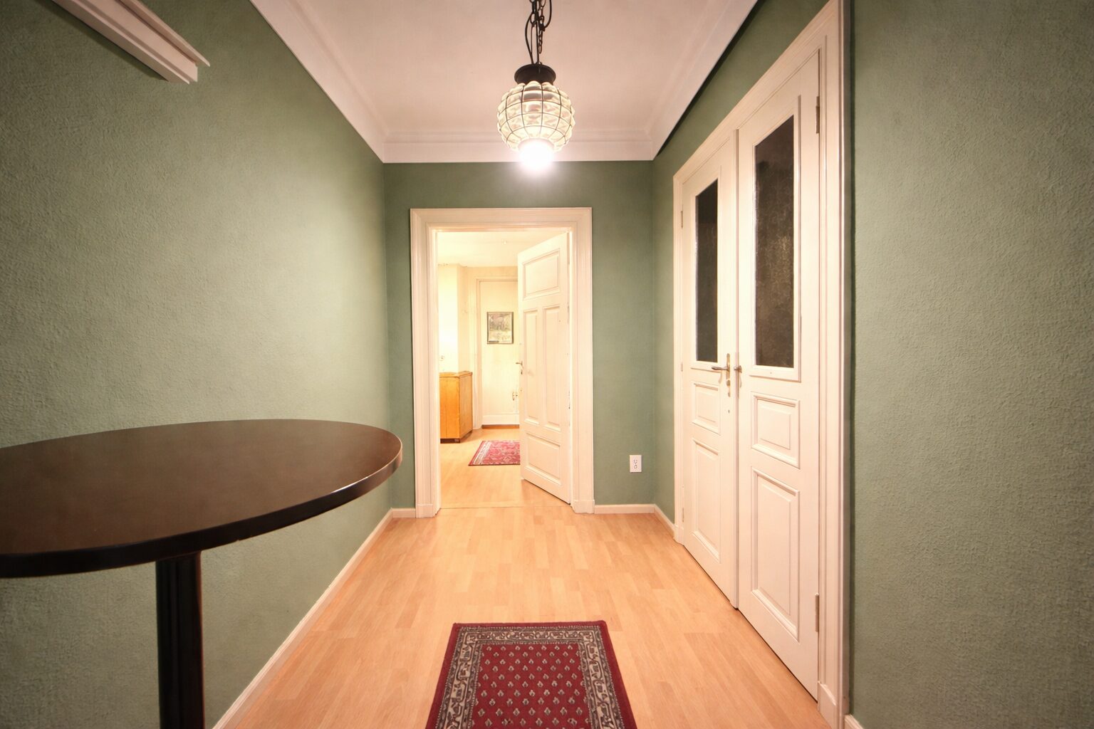 Green-walled hallway with a round wooden table on the left and white double doors on the right, lit by a hanging ceiling lamp; open doorway ahead reveals another room and rug.
